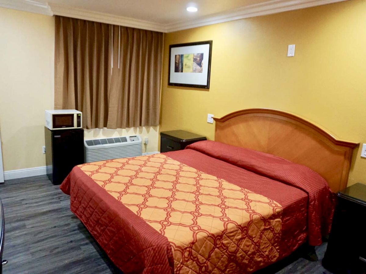 A hotel room with a double bed, red and yellow bedspread, bedside tables, microwave, air conditioner, framed picture, and closed curtains.