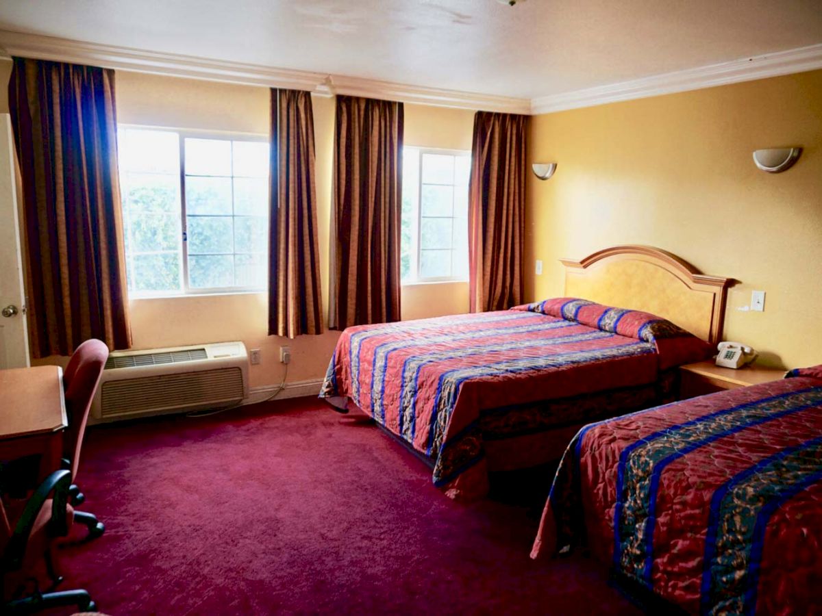 A hotel room with two double beds, a red carpet, a desk with a chair, two windows with curtains, an air conditioner, and a telephone.