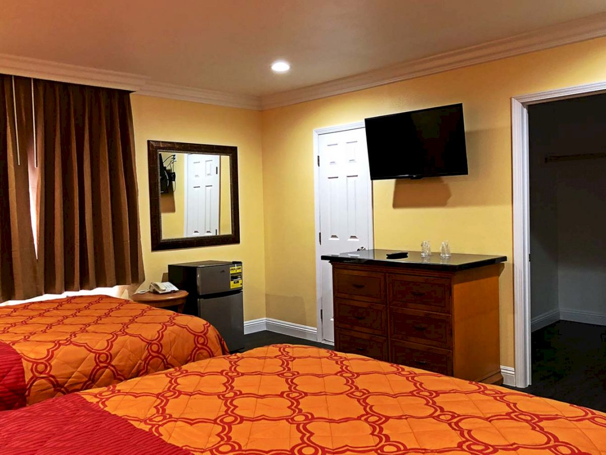 A hotel room features two beds, a TV mounted on the wall, a mini fridge, a dresser, and a mirror. The walls are painted yellow and the drapes are brown.