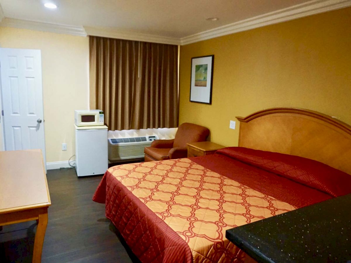 This image shows a hotel room with a double bed, desk, chair, mini-fridge, and microwave. The room has brown curtains and yellow walls.