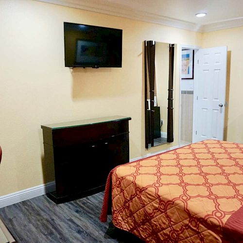 A modest hotel room with a bed, a wall-mounted TV, a microwave, a dresser, and a chair near a desk, featuring beige walls and wooden flooring.