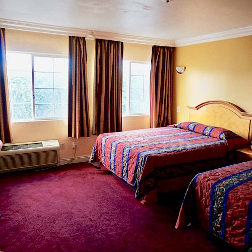 A hotel room with two beds, a desk, a chair, a phone, and brown curtains, illuminated by natural light from two windows.