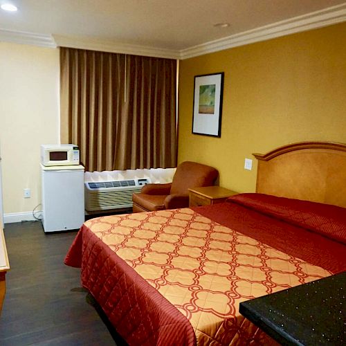 This image shows a hotel room with a bed, microwave, mini-fridge, chair, desk, air conditioner, and a framed picture on the wall.