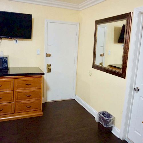 A room features a dresser with drawers, a microwave on top, a wall-mounted TV, a mirror, a door, and a trash can. The walls are painted light yellow.