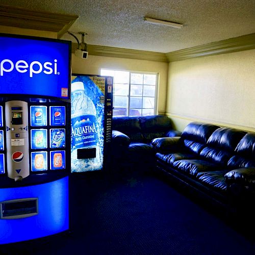 The image shows a room with a Pepsi vending machine and a second vending machine, along with dark leather sofas arranged near a window.