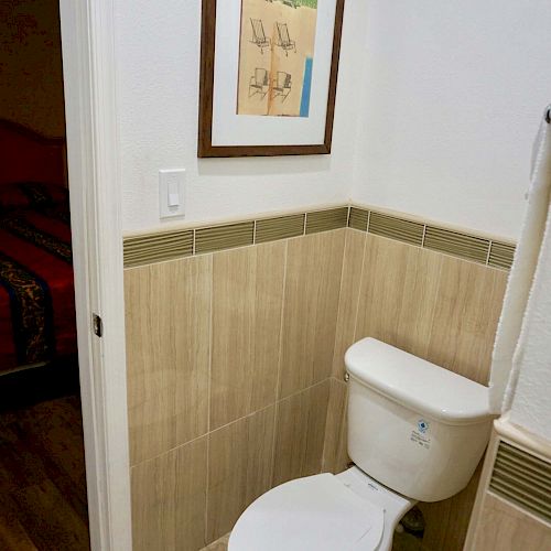 This image shows a small bathroom with a toilet and wall art above it. The wall art depicts a beach scene framed in brown wood.