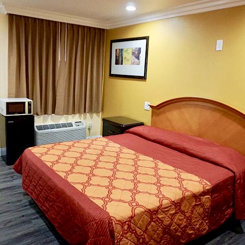 This image shows a bedroom with a double bed, two nightstands, a framed picture, a window with brown curtains, a microwave, and an air conditioner.