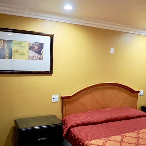 A neatly made bed with a red bedspread, flanked by two bedside tables, yellow walls, and framed artwork above the headboard adorn the room.