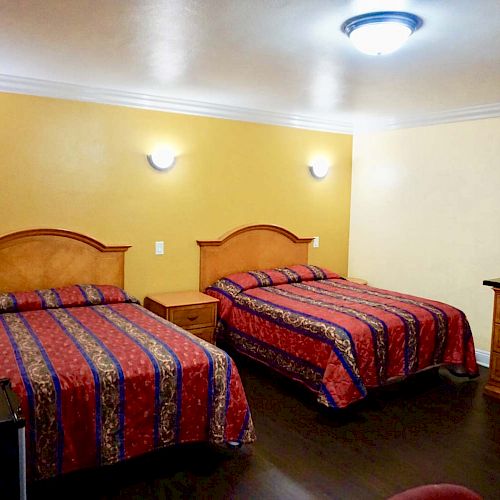 A hotel room with two double beds, a flat-screen TV on a wooden dresser, and colorful bedspreads. The walls are painted yellow and beige.