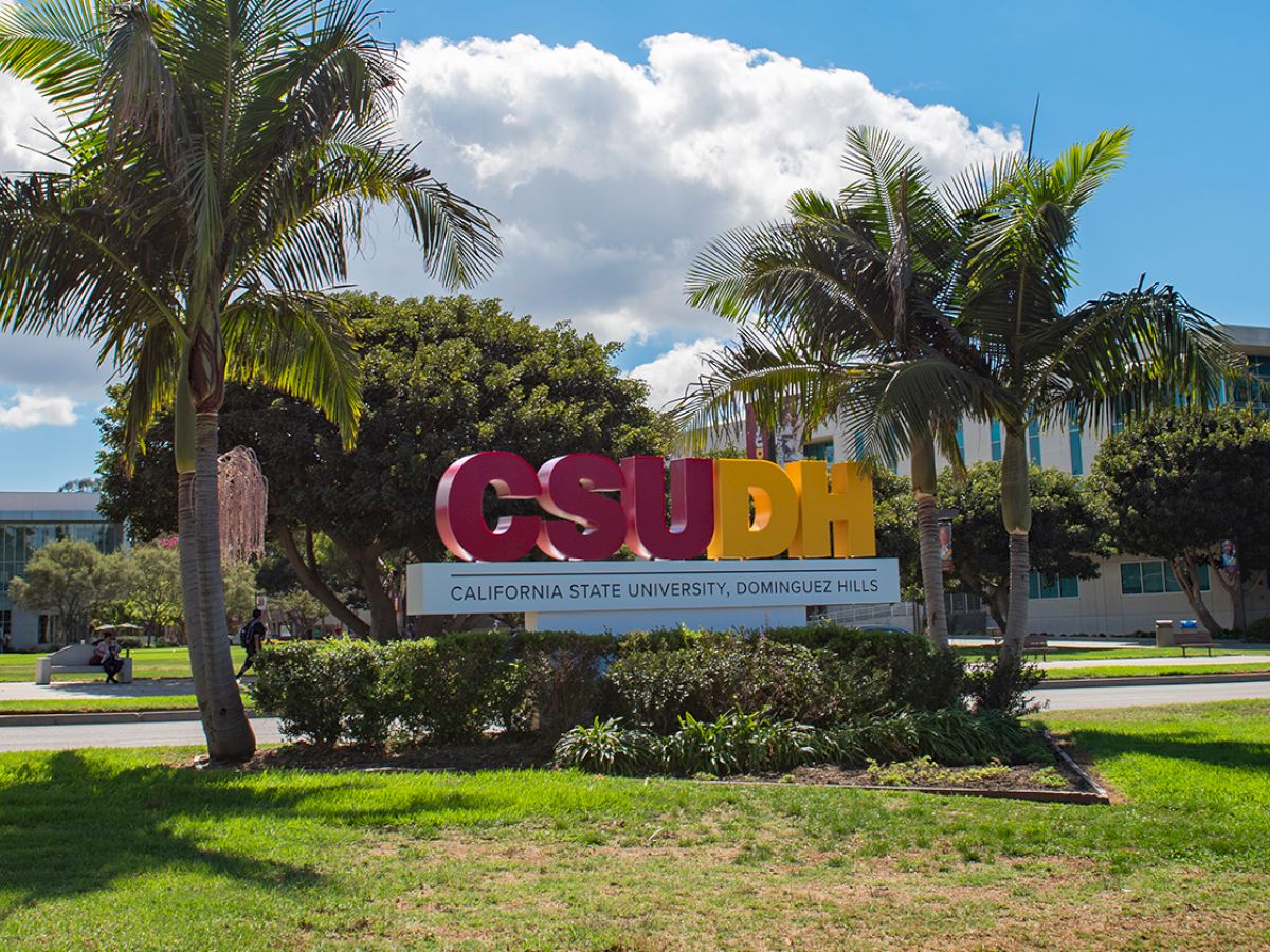 The image shows the entrance sign of California State University, Dominguez Hills (CSUDH), surrounded by palm trees and greenery ending the sentence.