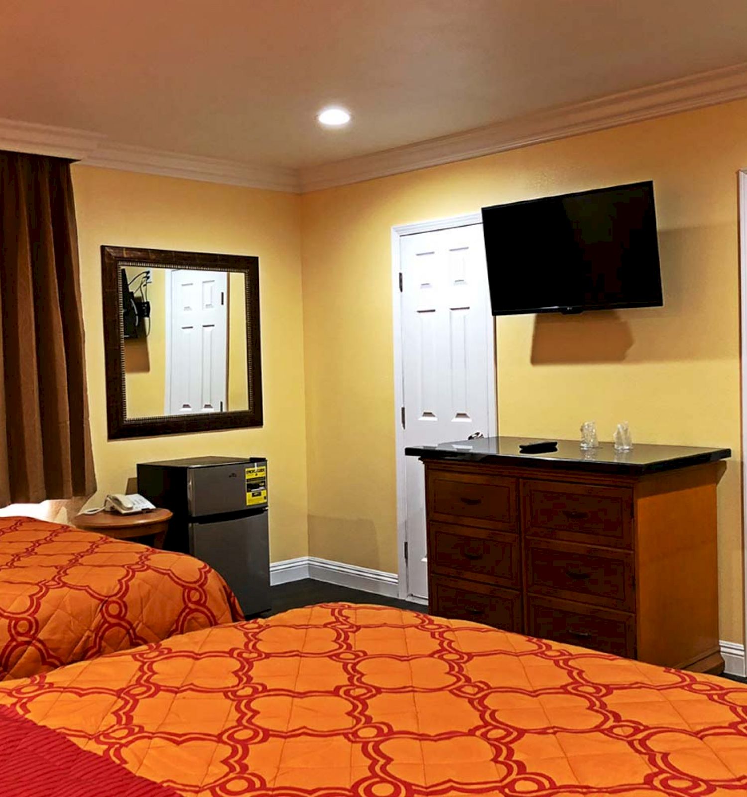 The image shows a hotel room with two red and orange beds, a wall-mounted TV, a mirror, a mini fridge, and an open closet door.