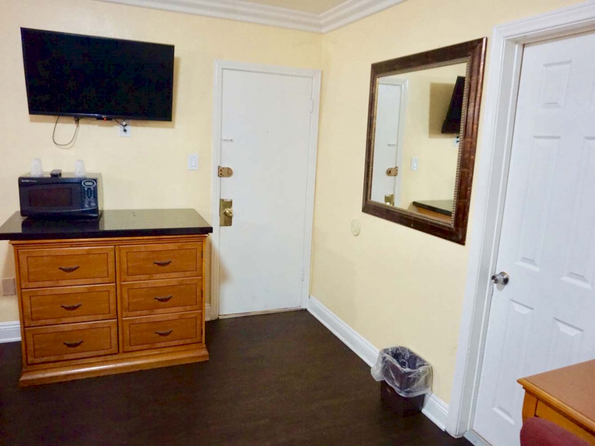 The image shows a room with a TV mounted on the wall, a wooden dresser with a microwave on top, a mirror, a door, and a small waste bin.