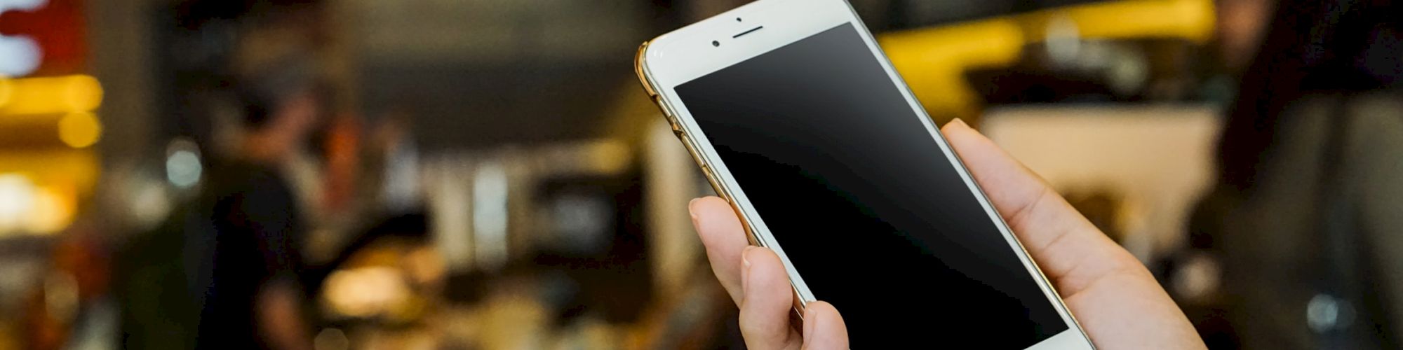 A hand holding a smartphone with a blank screen in a blurred background of what appears to be a café or restaurant setting.