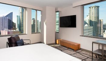The image shows a modern room with large windows, a bed, a chair, a wall-mounted TV, and a city view outside the windows.