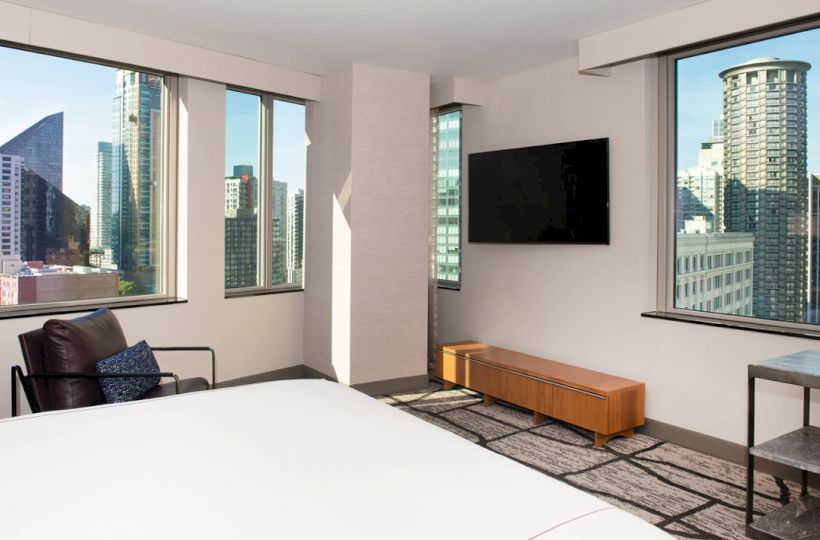 The image shows a modern room with large windows, a bed, a chair, a wall-mounted TV, and a city view outside the windows.