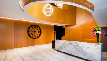 This image features a modern reception area with a marble desk, wooden wall, spiral staircase, large wall clock, and decorative lighting fixtures.