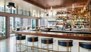 A modern bar area with a long counter, five blue bar stools, and a fully stocked liquor shelf, with large windows providing a city view.