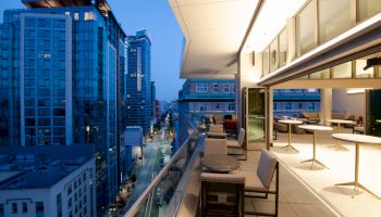 The image shows a modern cityscape at dusk with tall buildings and a balcony featuring outdoor seating and tables, overlooking a street.
