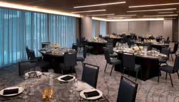 A banquet hall with round tables set with black napkins, wine glasses, and flower centerpieces. Soft lighting and large windows with blue curtains.