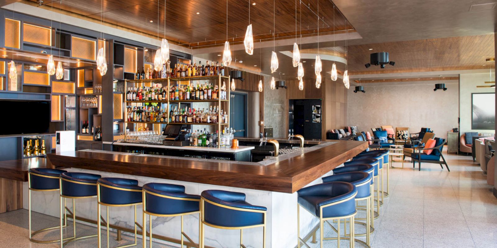 A modern bar with blue bar stools, a variety of bottles on shelves, hanging pendant lights, and a lounge area in the background.