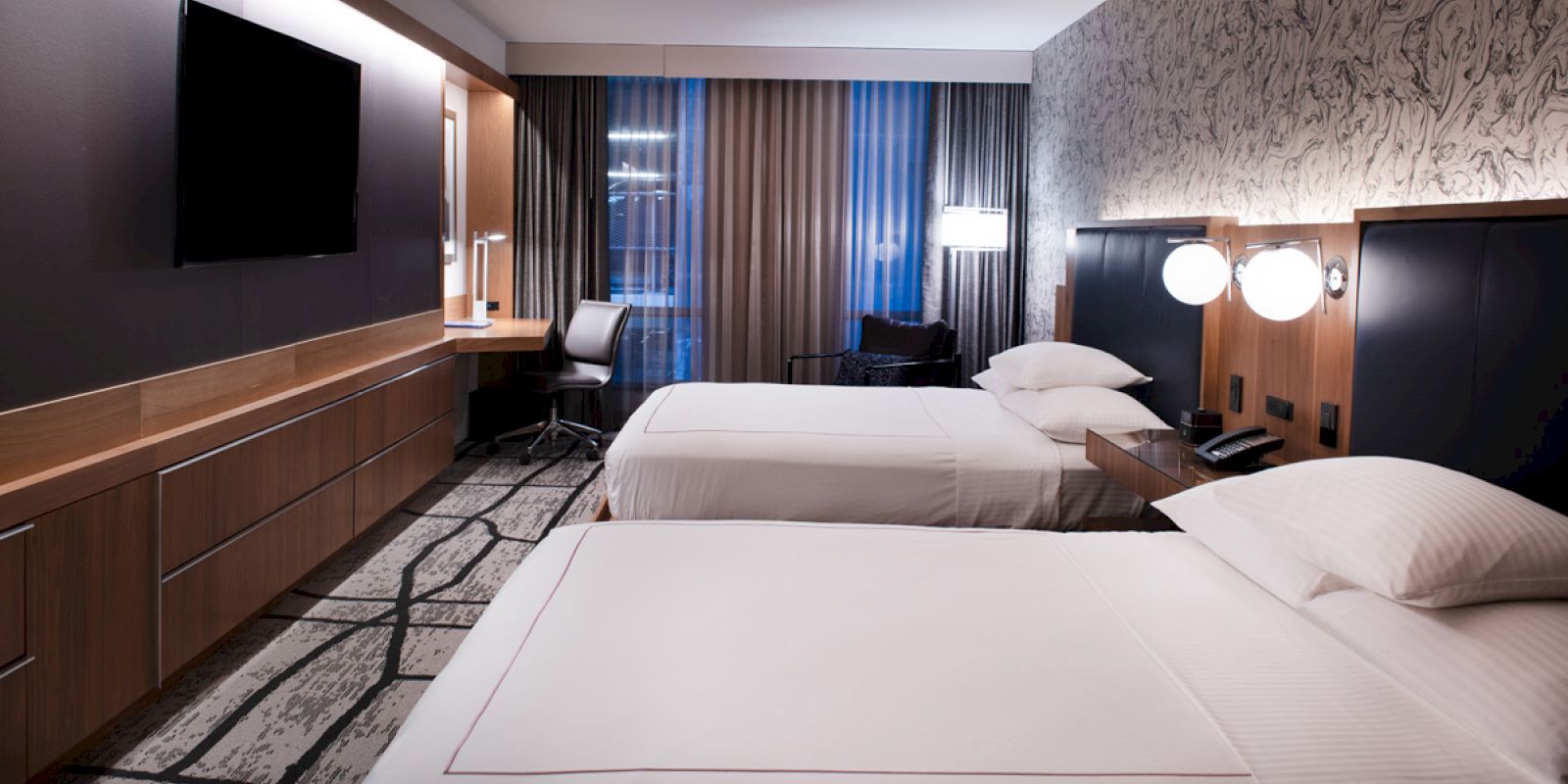 The image shows a modern hotel room with two neatly made beds, a wall-mounted TV, a desk with a chair, and large windows with closed curtains.