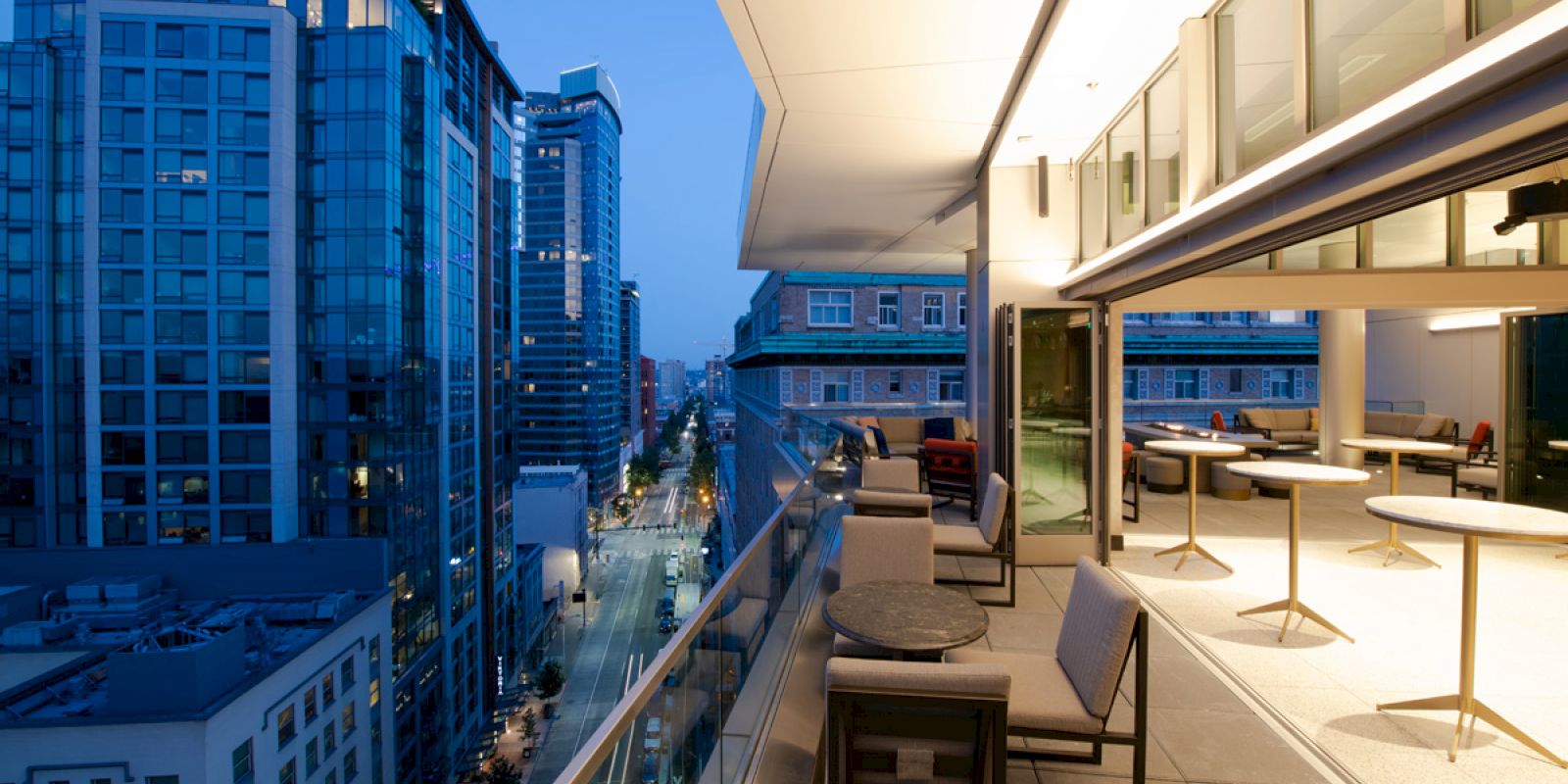 A city view at dusk from a modern balcony featuring outdoor seating and tables, with tall buildings and streetlights illuminating the scene.
