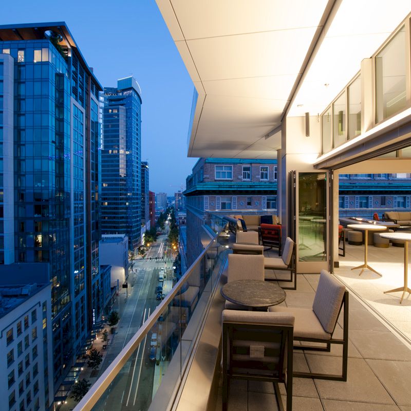 Modern cityscape during twilight with a balcony overlooking high-rise buildings; the balcony features seating and tables, offering cozy ambiance.