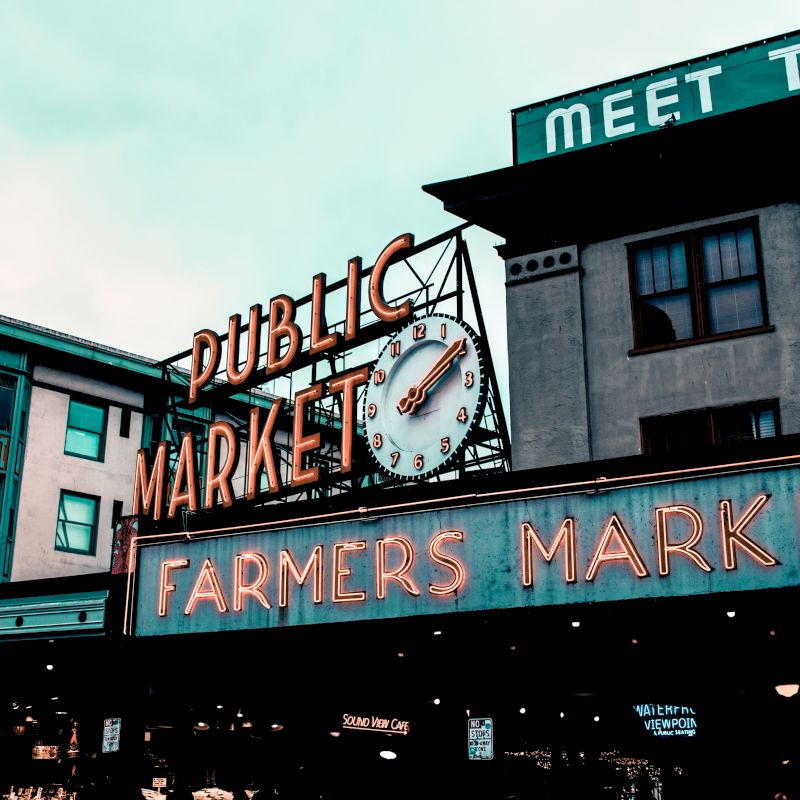 A neon sign reading "Public Market Center" and "Farmers Market" is shown, with a clock above. Buildings surround the market area.
