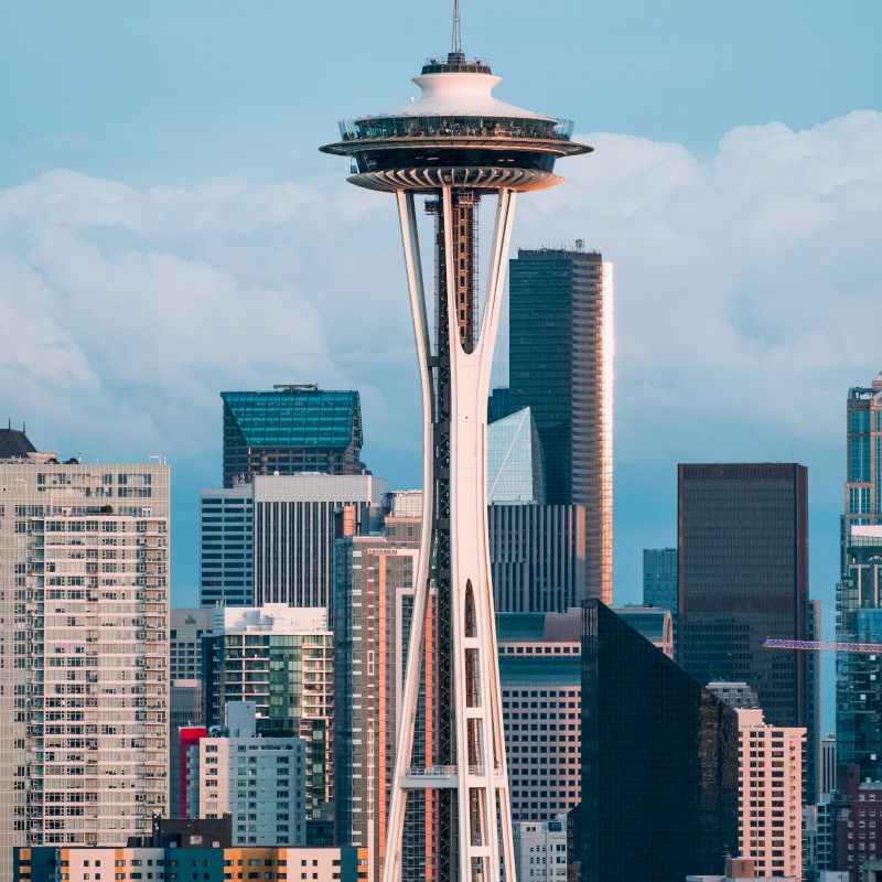 The image shows the Space Needle, a prominent observation tower in Seattle, with surrounding city buildings under a blue sky, and clouds in the background.