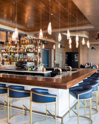 The image shows a modern bar with a wooden counter, blue bar stools, hanging lights, and a fully stocked liquor shelf in a spacious, stylish setting.