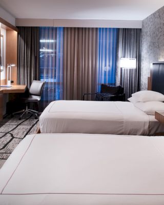The image shows a modern hotel room with two neatly made beds, a flat-screen TV, a desk and chair, and stylish lighting and wall decor.