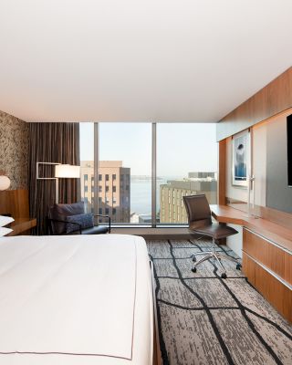 The image shows a modern hotel room with a large bed, a desk with a chair, a flat-screen TV, and a window with a city and water view.