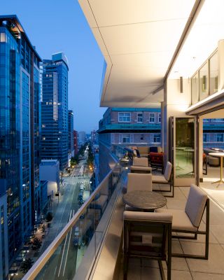 A modern balcony with seating and tables overlooks a city street lined with tall buildings at twilight, creating a sophisticated urban atmosphere.