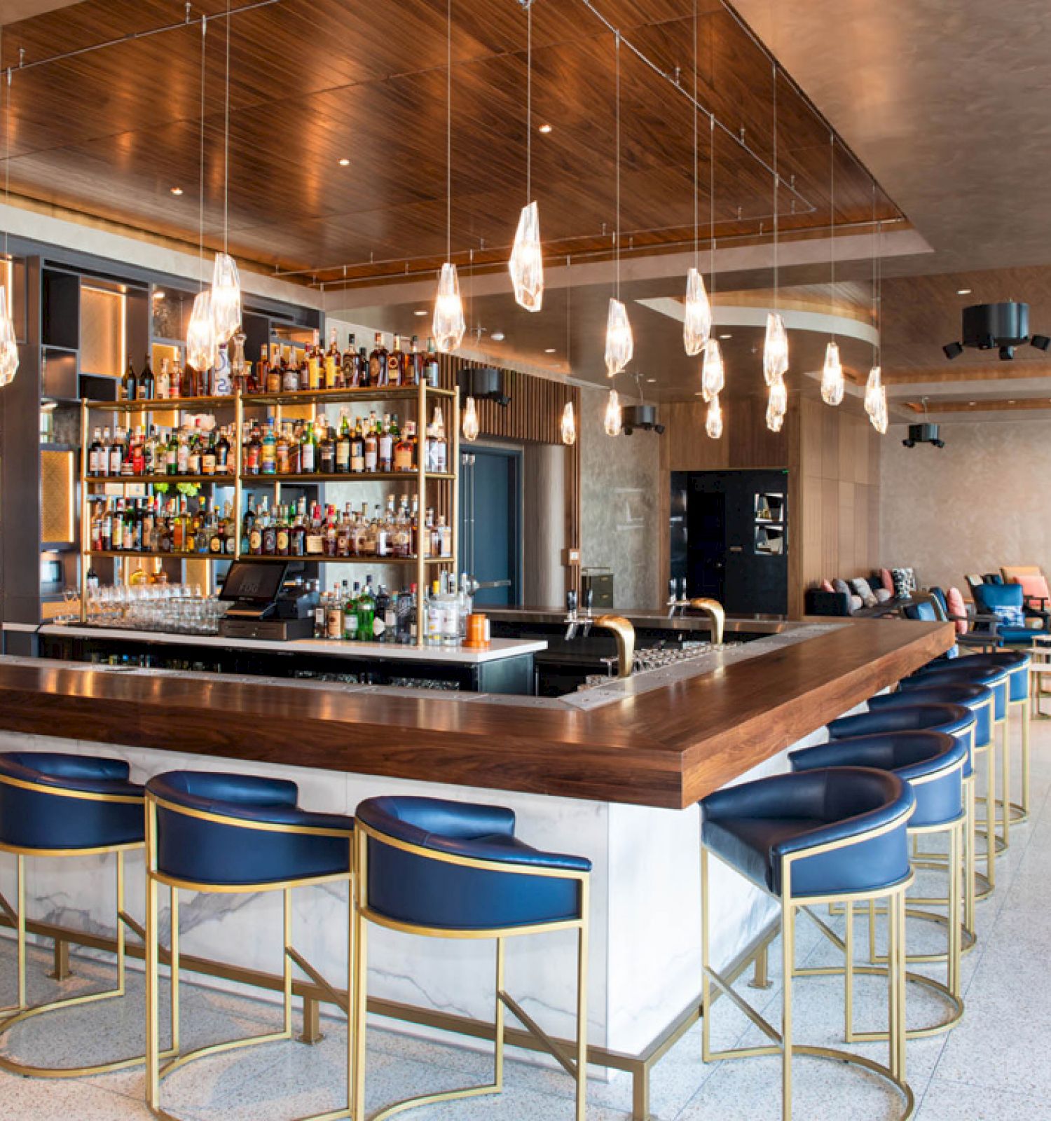 The image shows a modern bar with blue chairs, a wooden counter, hanging lights, and a fully stocked liquor shelf.