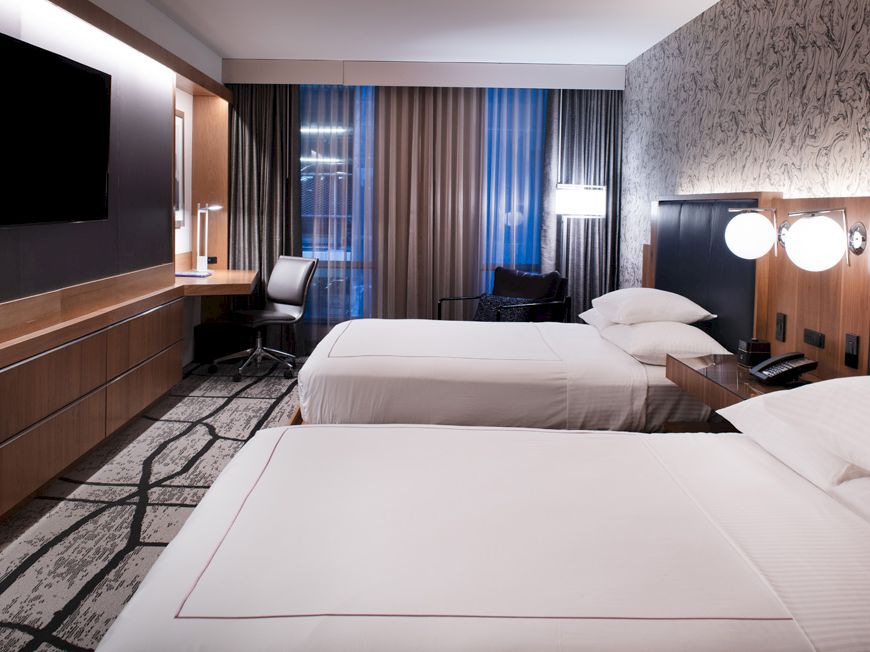 A hotel room with two neatly made beds, a TV on the wall, a desk, and modern decor featuring a patterned wall and large windows.
