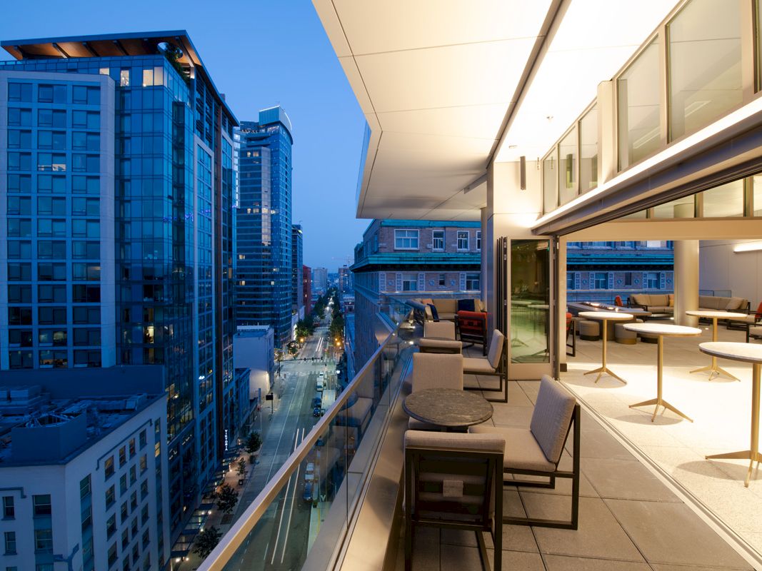 A modern, high-rise balcony at dusk with outdoor seating and tables overlooks a city street and adjacent office buildings.