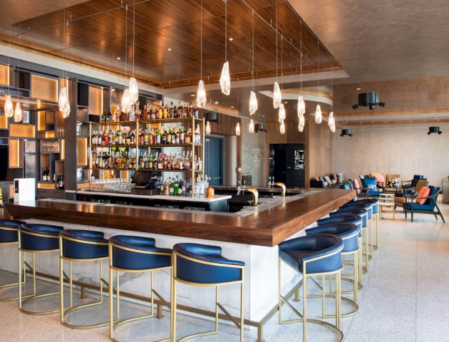 The image shows a modern bar with high stools, a well-stocked liquor shelf, and stylish lighting. There is also seating in the background.