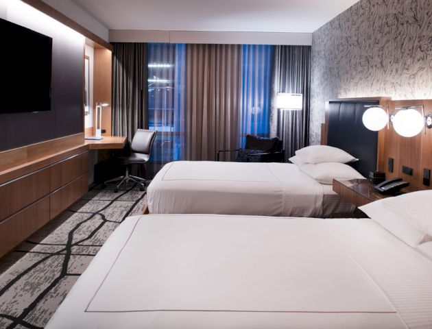 A modern hotel room with two beds, a flat-screen TV, a desk, and a window with curtains. The decor is contemporary with muted tones.