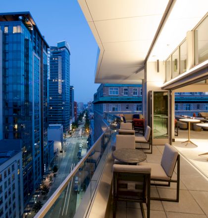 A modern high-rise balcony at dusk with outdoor seating overlooks a cityscape of tall buildings and a street filled with lights.