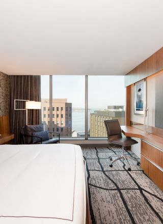 This image shows a modern hotel room with a large bed, a flat-screen TV, a desk, and a chair, featuring a city view through the window.