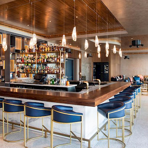 The image shows a modern bar with a wooden countertop, several bar stools, hanging lights, and a well-stocked alcohol shelf, along with a seating area.