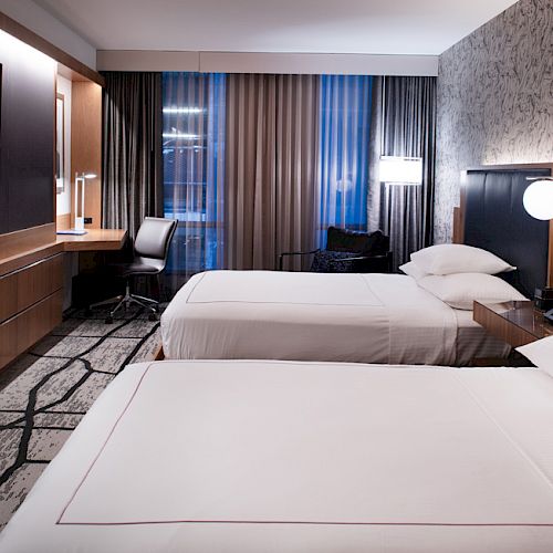 The image shows a modern hotel room with two neatly-made beds, a wall-mounted TV, a desk with a chair, and stylish decor with soft lighting.