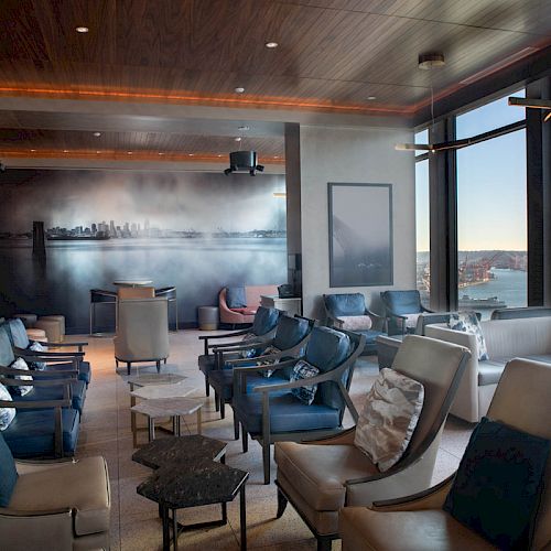 This image shows a modern lounge with comfortable seating, large windows offering a sea view, and a mural depicting a skyline on the back wall.