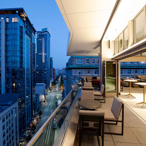 A modern rooftop terrace with seating overlooks a city street lined with tall buildings at dusk. The indoor area is well-lit with tables.