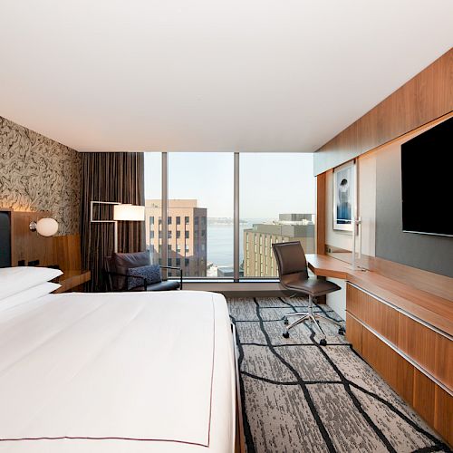 A modern hotel room with a large bed, wall-mounted TV, desk, chair, and floor-to-ceiling windows showing city buildings and a water view outside.