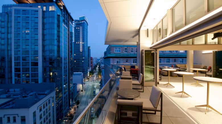 A modern cityscape at dusk with tall buildings and a view from a stylish terrace featuring outdoor seating and tables, alongside a street below.