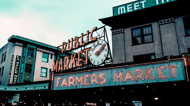 The image shows the iconic neon signs of a "Public Market" and "Farmers Market" with a clock, likely at a popular market location.