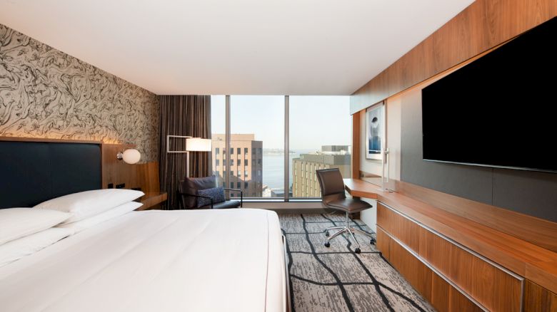 A modern hotel room with a large bed, wall-mounted TV, desk, chair, and a window offering a city view. Decor includes patterned wallpaper.