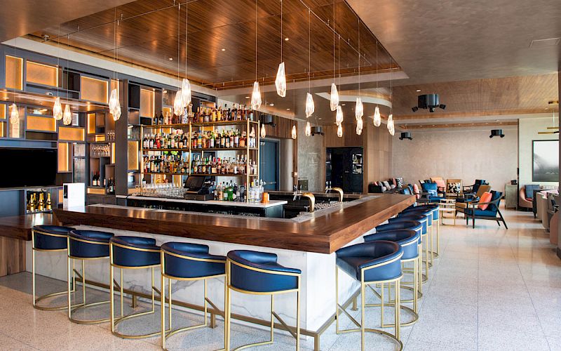 This image shows a modern bar with blue chairs, a stocked bar, pendant lights, and a lounge area in the background.