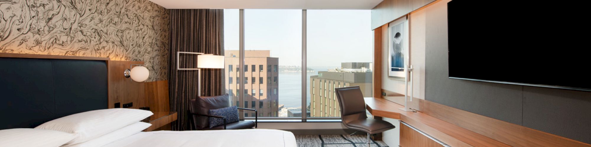 A modern hotel room with a large bed, wall-mounted TV, desk with a chair, large windows with a city view, and contemporary decor.